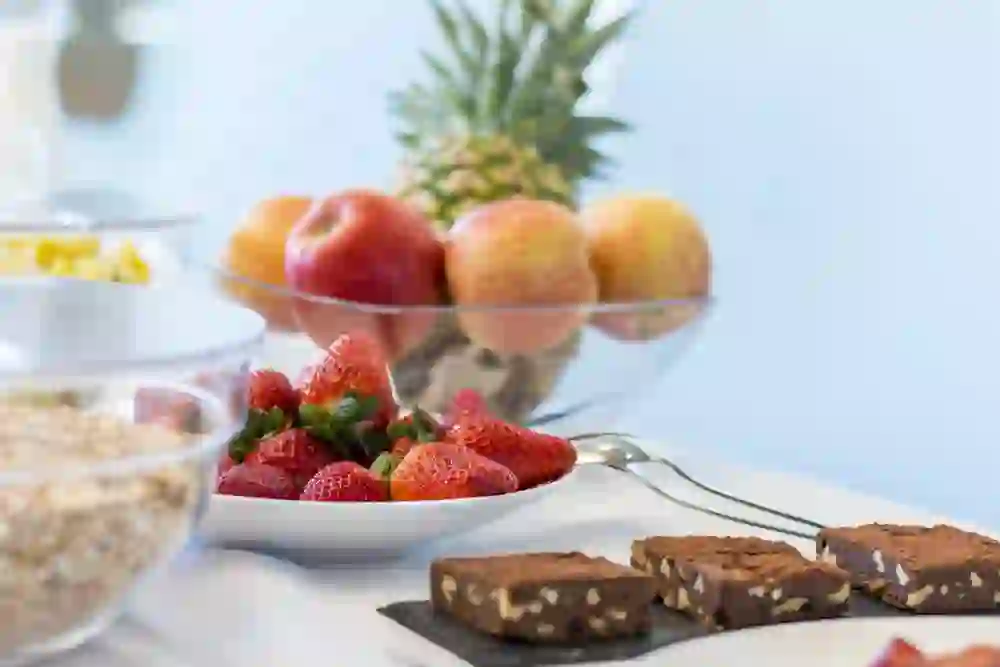 Auf dem Foto ist ein Teil eines Frühstücksbuffets zu sehen. Im Vordergrund sind frische Erdbeeren auf einem Teller und Schokoladenbrownies auf einem Schieferplättchen angerichtet. Dahinter befinden sich Schalen mit Müsli und eine Glasschale mit Äpfeln, während eine Ananas das farbenfrohe Arrangement komplettiert. Die Präsentation ist ansprechend und wirkt einladend.