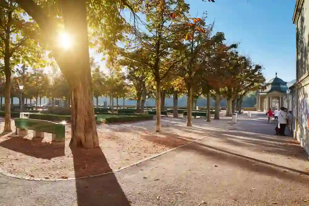 L'image présente la plate-forme de la cathédrale de Berne sous un soleil radieux qui se brise à travers les feuilles des arbres, créant une ambiance automnale. Les gens se promènent en toute décontraction, profitant de la chaleur et du jeu d'ombre et de lumière qui créent une atmosphère paisible et pleine de joie de vivre.