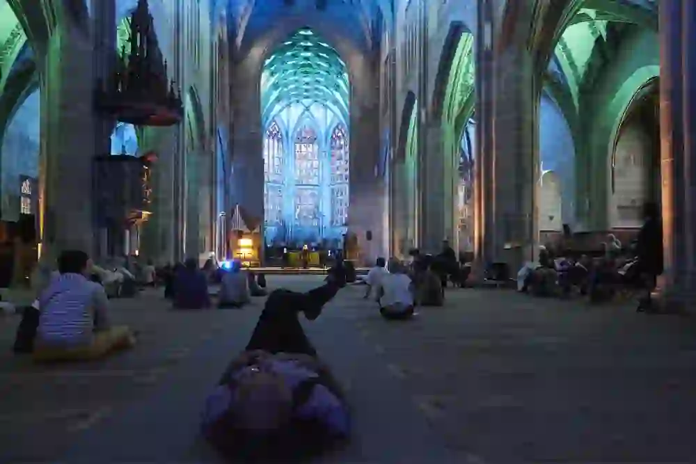 In the subdued lighting of the French Church in Bern, surrounded by the majestic arches and stained glass windows, the audience gathers for a concert at the music festival. The atmosphere is relaxed and reverent, with people lying or sitting on the floor, completely captivated by the sacred atmosphere and the preparatory sounds of the music.