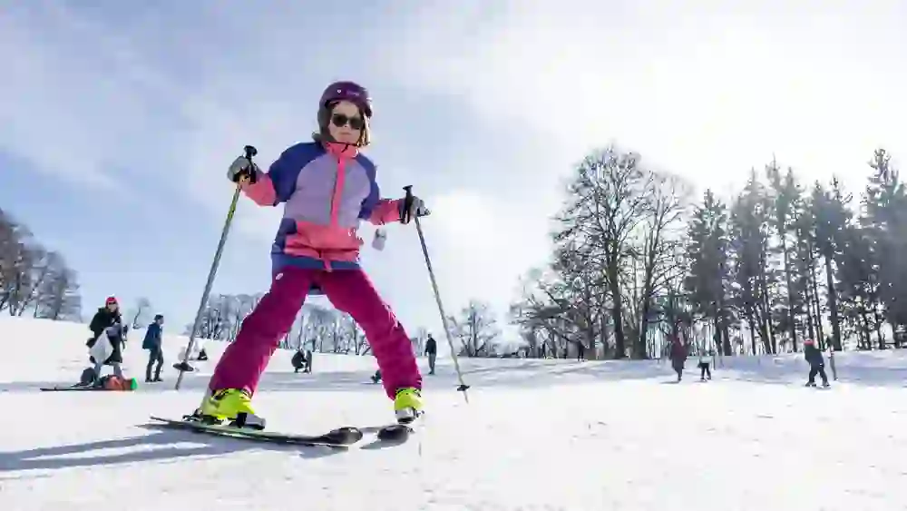 Un enfant dévale la pente à ski, concentré, les bâtons bien en main. En arrière-plan, d'autres amateurs de sports d'hiver s'entraînent. L'atmosphère est fière, ludique et pleine d'enthousiasme enfantin.