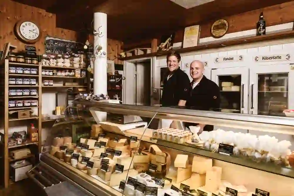 A l'intérieur de la "Chäshütte", deux personnes sourient derrière un comptoir rempli de spécialités fromagères, ce qui donne une atmosphère chaleureuse et accueillante.