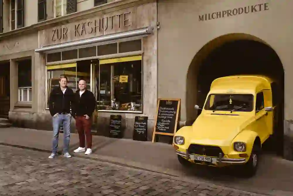 Vor der "Chäshütte" stehen zwei lächelnde Personen neben einem gelben Lieferwagen, was eine freundliche und willkommene Stimmung vermittelt.
