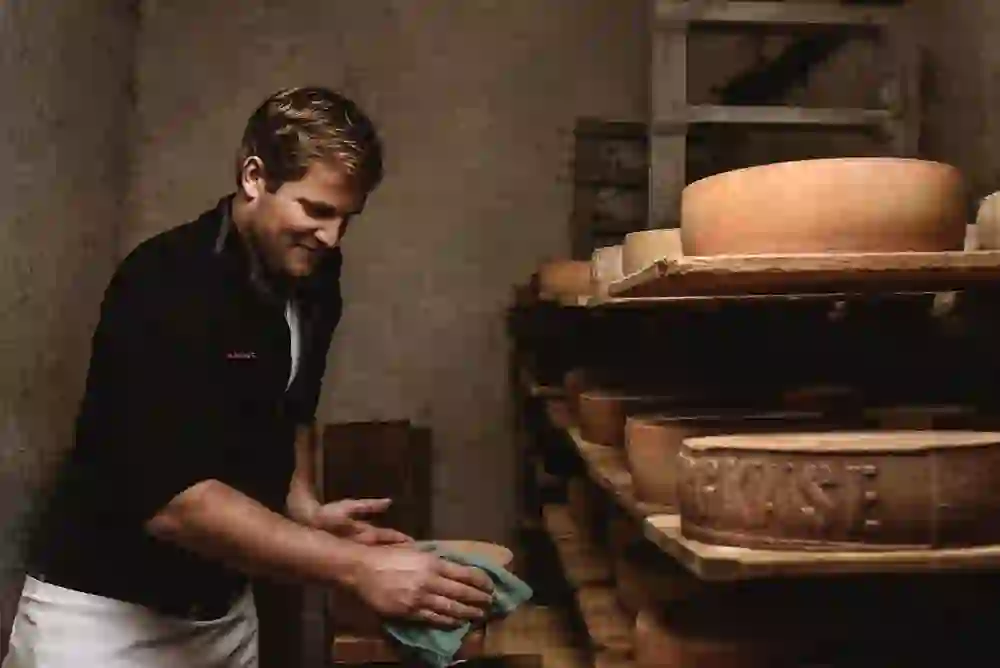 A cheesemaker carefully tends large wheels in a cheese cellar, surrounded by an atmosphere of traditional craftsmanship and dedication.