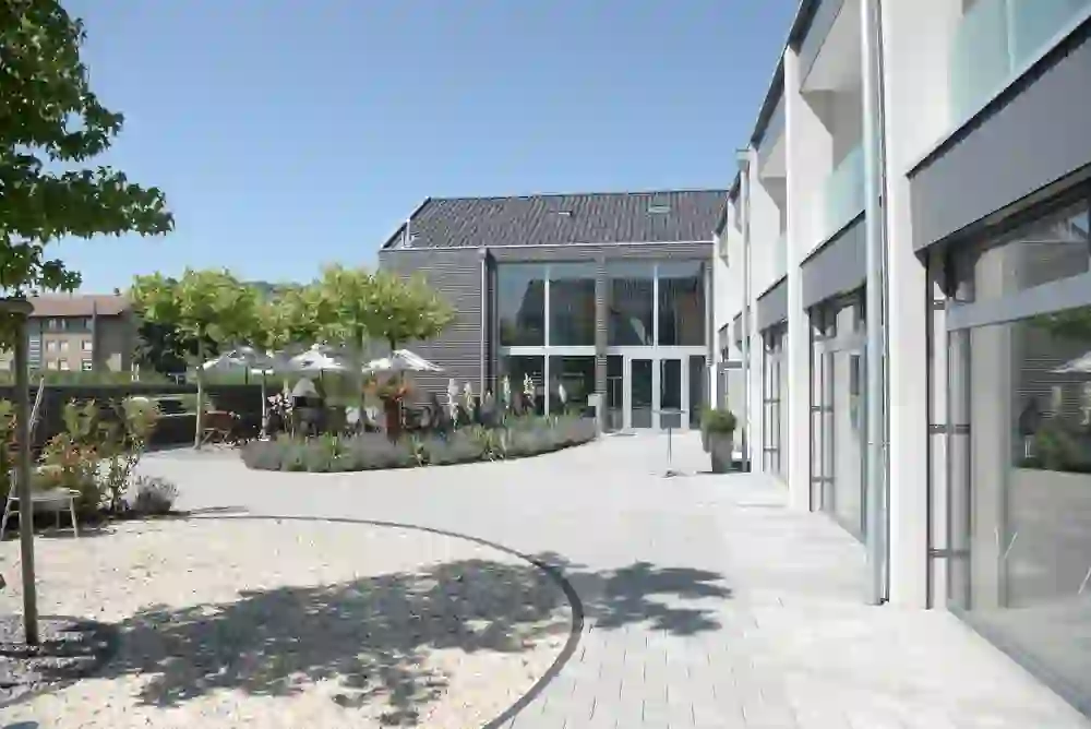 Sur la photo, on peut voir un bâtiment moderne avec une grande façade vitrée et une allée pavée, entouré d'un jardin bien entretenu. Le décor est accueillant et représente une architecture moderne dans un environnement calme du QCM Campus Belp.