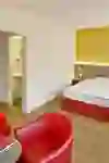 La chambre d'hôtel combine des tons chauds de jaune et de rouge avec des éléments en bois et crée une atmosphère confortable et accueillante. Deux fauteuils rouges invitent à la détente.