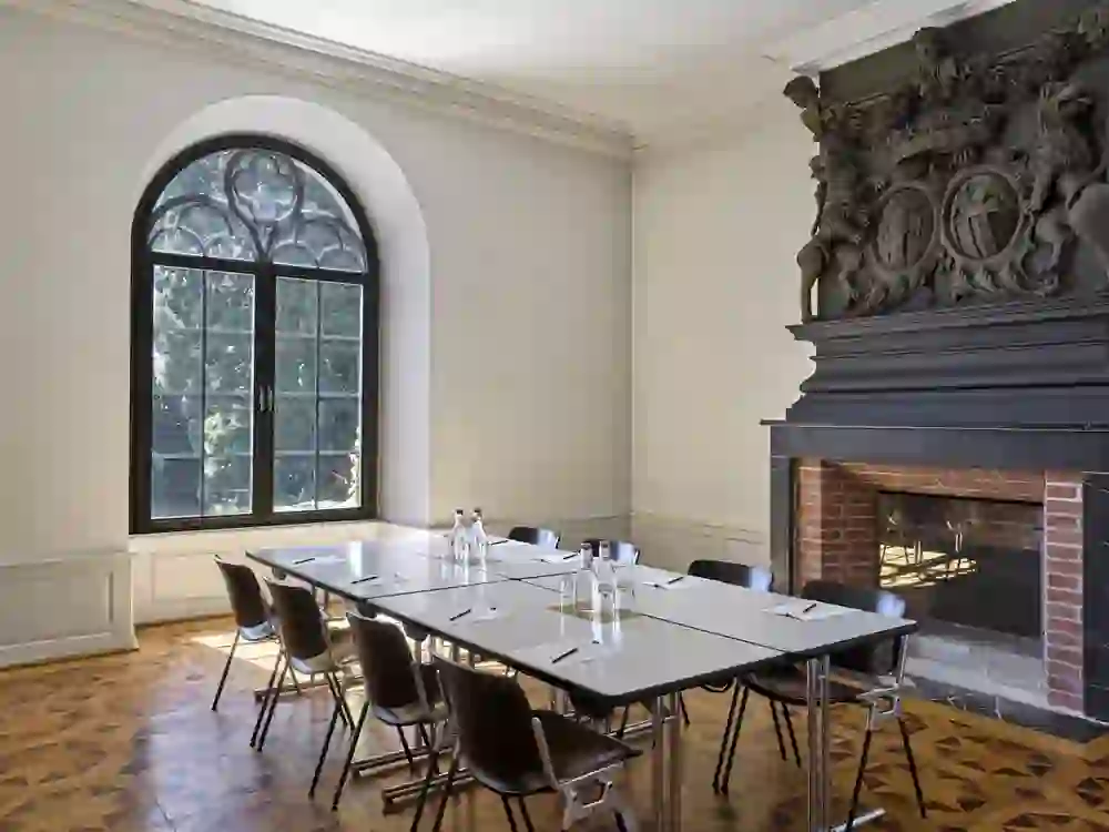 Der Raum im Schloss Münchenwiler, mit einem eleganten Konferenztisch, ist durch ein großes Bogenfenster natürlich beleuchtet. Die ruhige und stilvolle Atmosphäre wird durch den historischen Kamin betont, ideal für formelle Besprechungen.