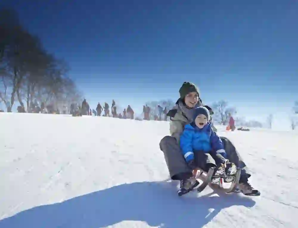 Eine fröhliche Schlittenfahrt im Gurtenpark: Ein Erwachsener und ein kleines Kind, dick eingepackt gegen die Kälte, sausen mit strahlenden Gesichtern den verschneiten Hang hinunter, pure Freude im Winterwunderland.