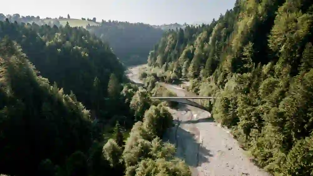 Une perspective de drone révèle l'étroit pont Maillart, qui relie deux côtés d'une vallée boisée au-dessus d'un cours d'eau caillouteux. Les forêts denses qui l'entourent forment un mur vert naturel qui souligne le caractère sauvage et la beauté intacte de la région.