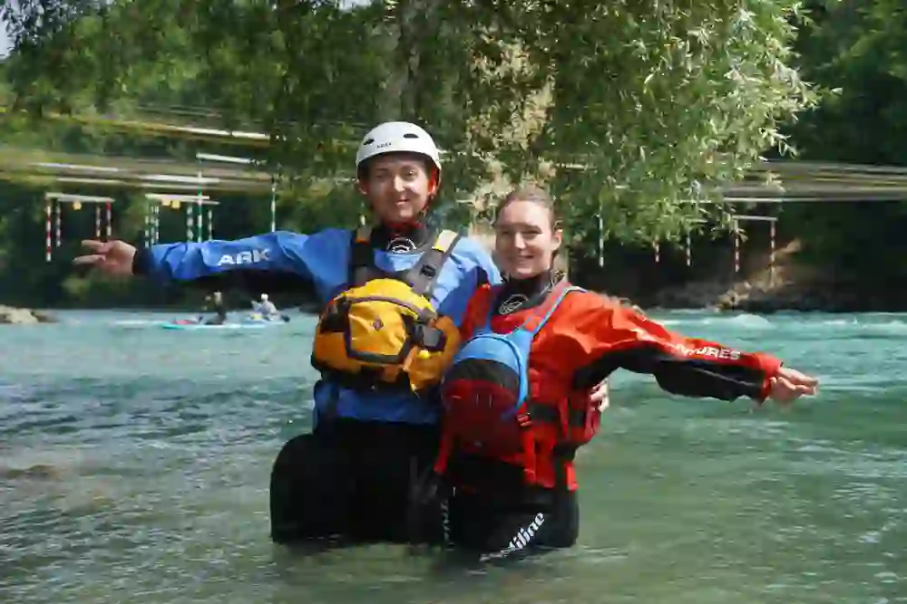 L'immagine mostra due giovani che si trovano insieme in un fiume e sorridono all'obiettivo. Entrambi indossano mute colorate (rosse e blu), giubbotti di salvataggio e caschi: il loro equipaggiamento indica chiaramente un'attività di acqua bianca o kayak. Sullo sfondo si vedono acque calme di colore turchese, altri canoisti e un percorso di slalom con pali di cancello sospesi, incorniciato da una vegetazione lussureggiante. L'atmosfera è vivace, sportiva e vicina alla natura: l'immagine irradia un senso di avventura e di spirito di squadra.