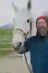 Das Bild zeigt einen lachenden Mann in blauer Jacke und roter Mütze, der neben einem weißen Pferd steht. Die friedliche ländliche Umgebung mit grünen Wiesen und Bergen im Hintergrund vermittelt eine warme, freundliche Stimmung und die Freude am Zusammensein mit dem Tier.