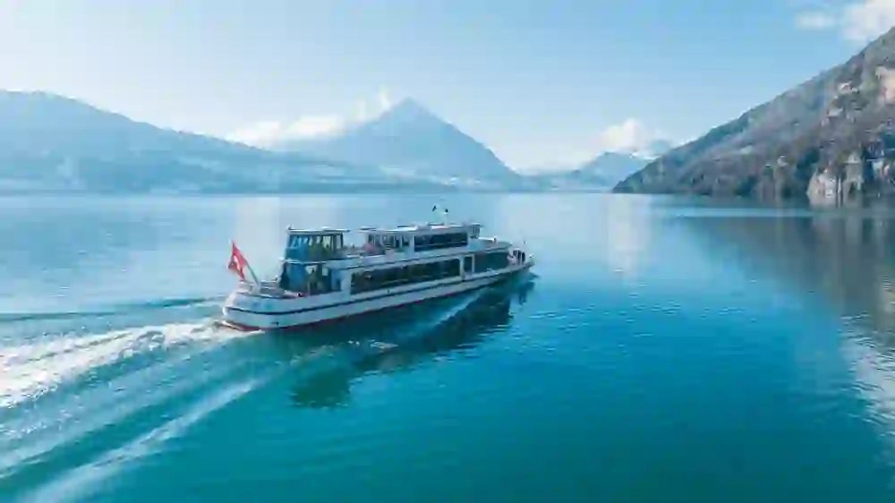 The picture captures the peaceful scenery of a winter cruise on Lake Thun. A boat glides gently over the calm waters, surrounded by a picturesque snowy landscape and an imposing mountain panorama under a clear sky.