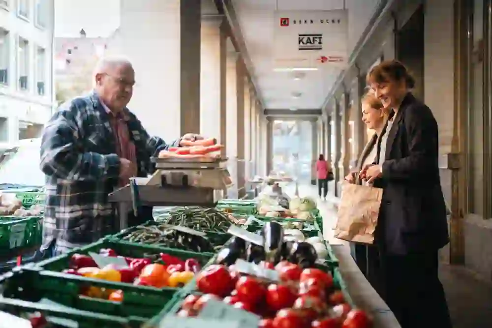 La foto mostra una vivace bancarella del mercato dove un simpatico venditore pesa le verdure mentre due donne, che sembrano allegre, fanno acquisti. L'atmosfera è vivace e calda.