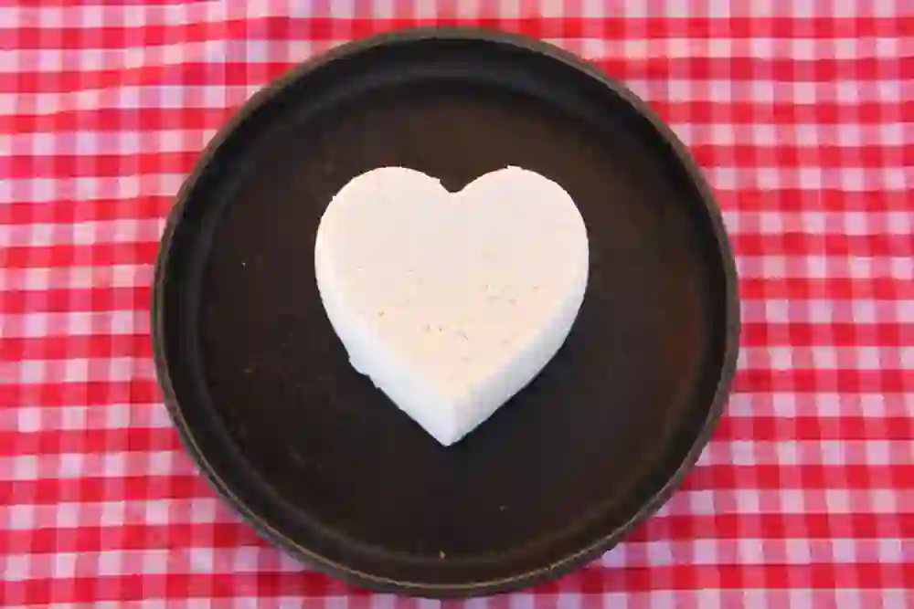 L'immagine irradia un'atmosfera rustica e accogliente. La crema di formaggio a forma di cuore su un piatto scuro, posto su una tovaglia a scacchi, trasmette un'atmosfera calda e rurale. Evoca associazioni di tradizione, amore per la natura e la piacevole semplicità del caseificio Emmental.