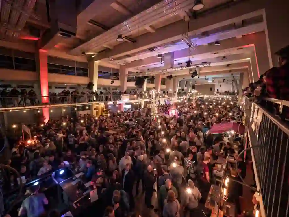 Auf dem Bild ist eine belebte Veranstaltungshalle mit einer Menschenmenge bei warmem, gemütlichem Licht zu sehen. Die Stimmung wirkt ausgelassen und gesellig.