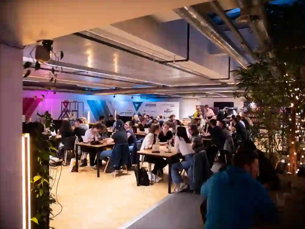 In der Erupt Gaming Lounge herrscht eine lebhafte Atmosphäre, wo Menschen um Tische versammelt sind, in angeregten Gesprächen und gemeinsamen Aktivitäten, umgeben von sanftem Licht und modernem Ambiente.