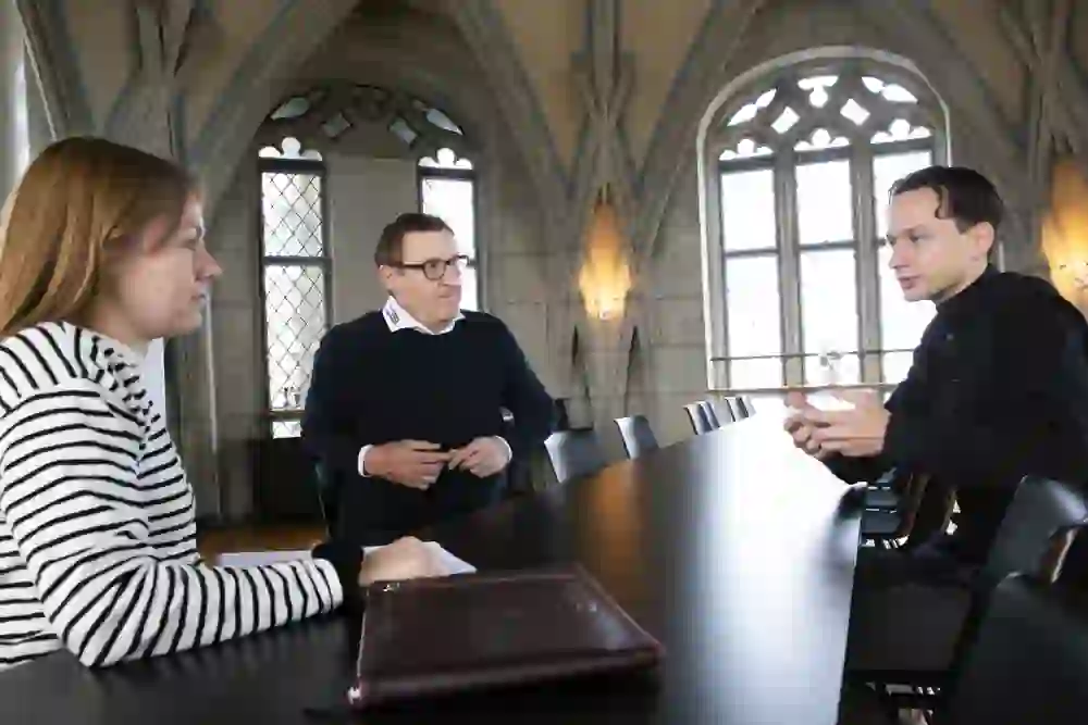 Dans une pièce aux fenêtres gothiques, une personne mène une conversation intense, soutenue par des gestes expressifs, tandis que les deux autres participants écoutent attentivement. L'atmosphère semble sérieuse et concentrée, éclairée par une lumière douce.