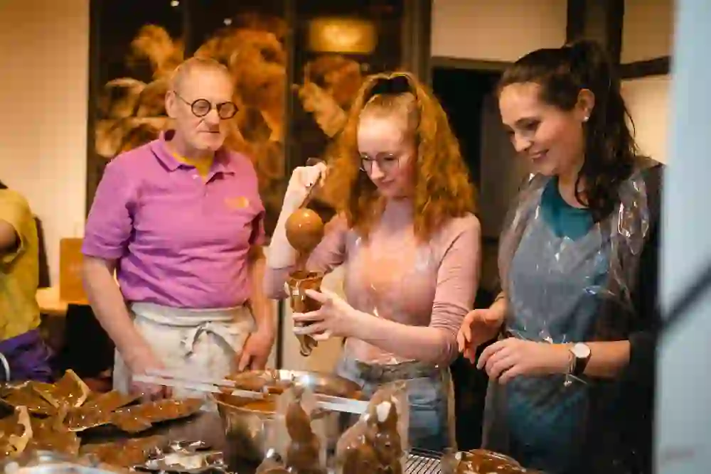 L'image montre un groupe de personnes qui coulent ensemble des lapins de Pâques en chocolat. Une jeune femme au centre verse du chocolat liquide d'une louche dans un moule à lapin, tandis qu'une autre femme observe en souriant. Un homme plus âgé, vêtu d'un tablier et d'un polo violet, se tient à côté d'elle et semble la soutenir ou la guider. La scène ressemble à un atelier de chocolatier ou de pâtissier, où les gens peuvent découvrir l'artisanat traditionnel de la fabrication du chocolat. Au premier plan, on peut voir des lapins en chocolat déjà finis ou à moitié finis ainsi que des éclats de chocolat et des outils. L'atmosphère chaleureuse indique qu'il s'agit d'un atelier créatif et gourmand pour Pâques.