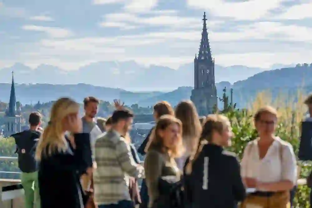 Das Bild zeigt Menschen im Gespräch auf einer Terrasse, mit Blick auf das Berner Münster und die Alpen im Hintergrund. Die Stimmung wirkt entspannt und gesellig, das Wetter ist klar und sonnig, was eine positive, offene Atmosphäre erzeugt.