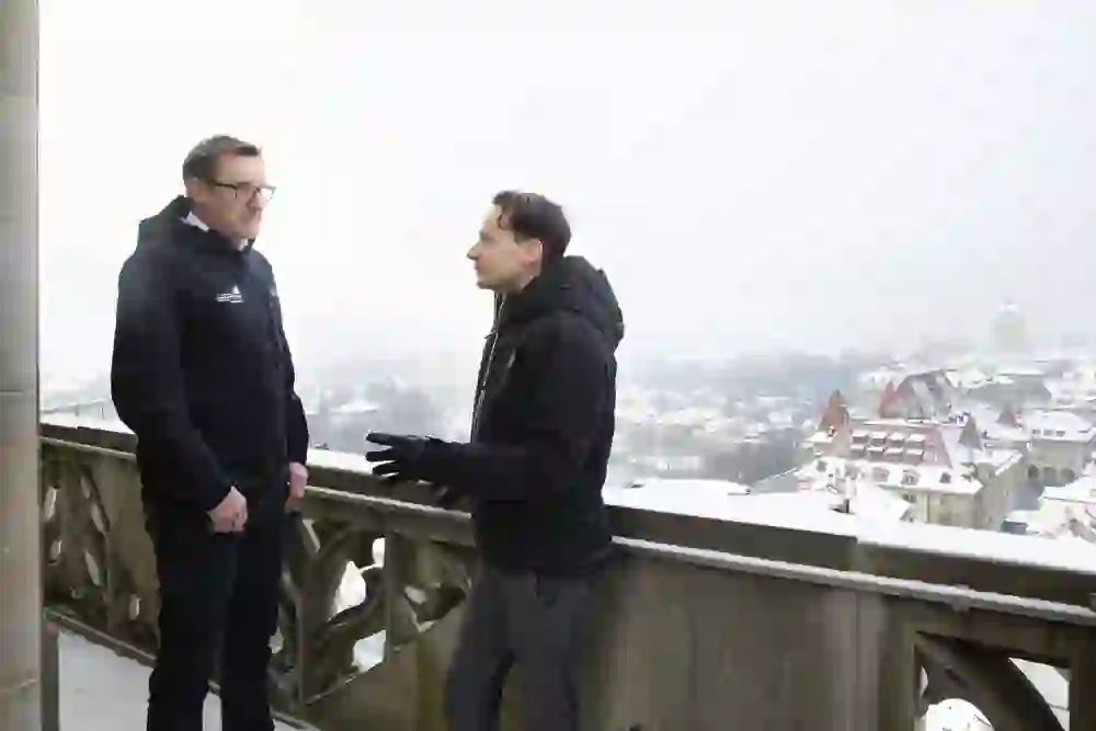 Deux hommes sont en train de discuter sur une terrasse recouverte de brume avec vue sur Berne enneigée. L'atmosphère est calme et pensive, ponctuée par l'échange animé entre les deux hommes.