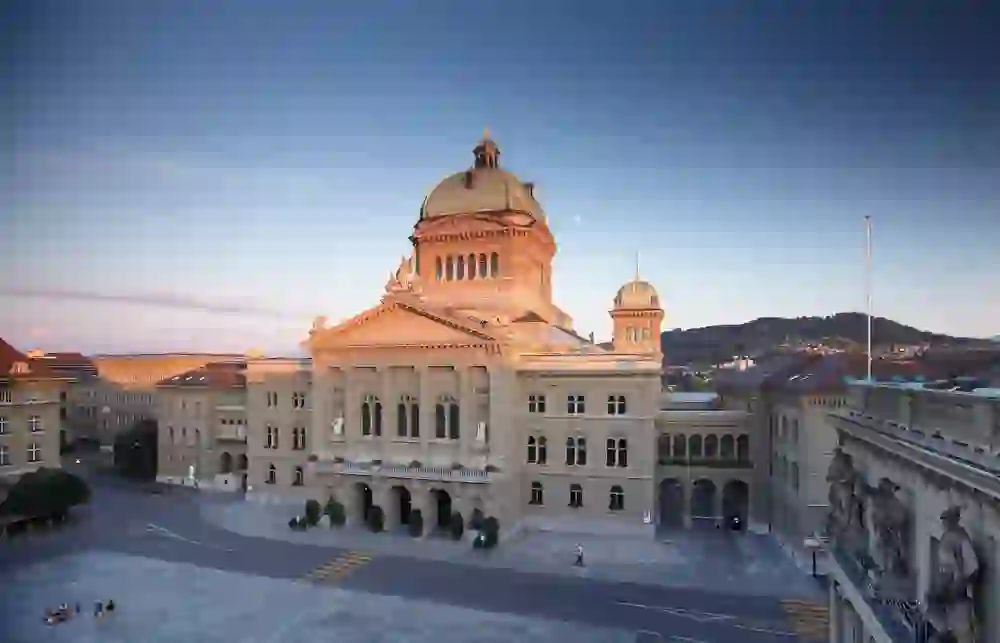 The Federal Palace in Bern is illuminated by the evening sun, creating a calm, dignified atmosphere as people enjoy the square in front of it.