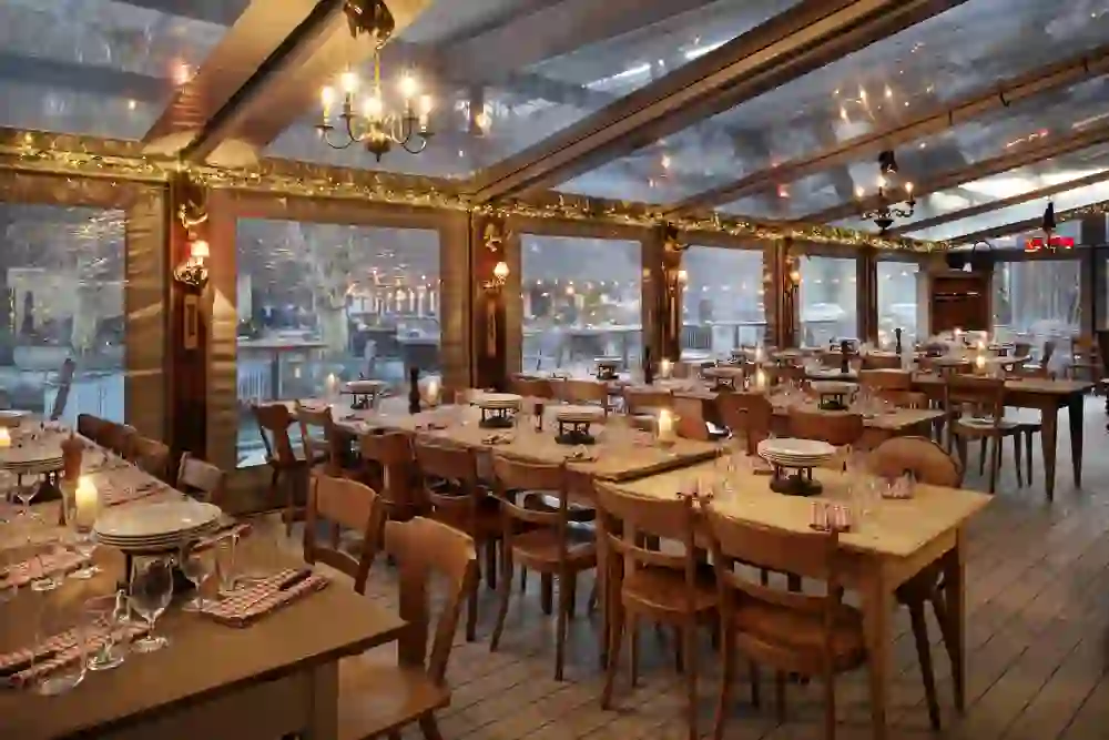 Gemütlich dekorierte Fondue-Hütte mit grossen Fenstern und durchsichtigem Dach, durch das man Bäume und Lichter von draußen erkennt. Die rustikalen Holztische sind festlich gedeckt mit Fondue-Rechauds, Kerzen, Gläsern und rot-weiß karierten Servietten. Warme Beleuchtung, Lichterketten und Kronleuchter sorgen für eine stimmungsvolle Atmosphäre.