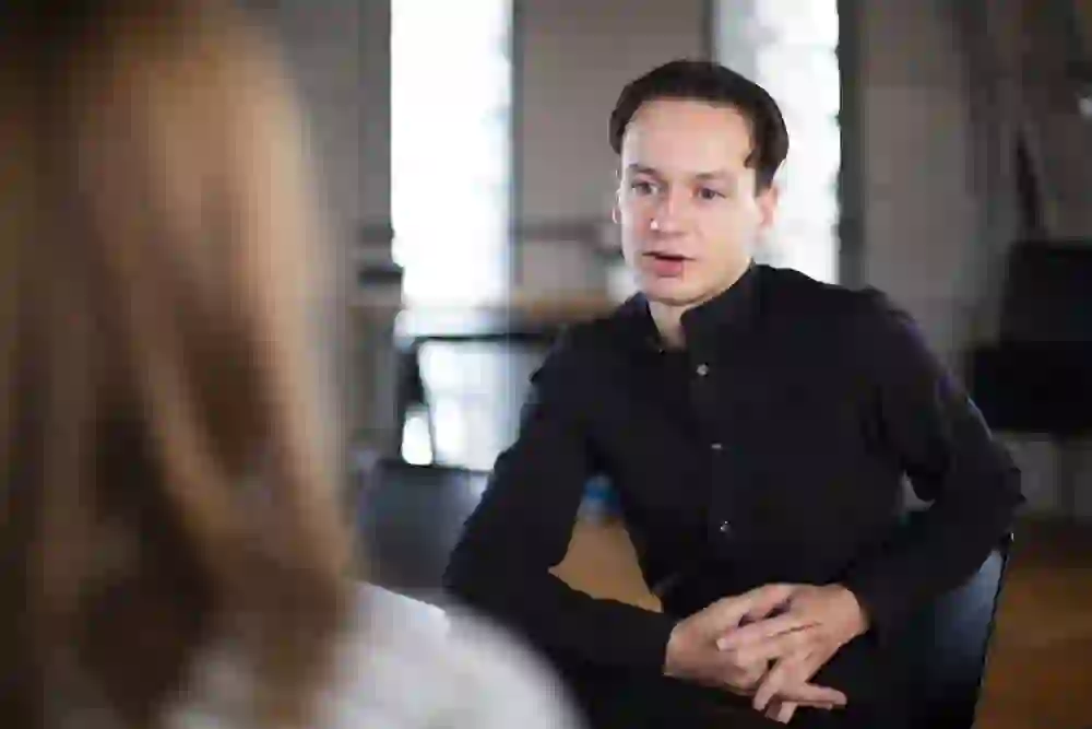 Un homme est assis en train de discuter, concentré et le regard attentif, ses mains soulignant l'intensité de l'échange. Un sentiment d'engagement et de sérieux emplit la pièce.