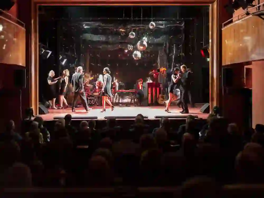 Das Foto zeigt eine lebhafte Aufführung im Casino Theater Burgdorf. Auf der Bühne befinden sich Musiker und Tänzer in eleganter Abendgarderobe. Das Publikum, eingetaucht in das warme Ambiente des Theatersaals, beobachtet die Szene mit sichtbarem Genuss. Die Stimmung ist festlich und ausgelassen.
