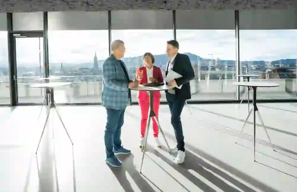 Drei Personen stehen in einem modernen, hell erleuchteten Raum mit großen Fenstern, die eine Aussicht auf eine Stadt bieten, und führen ein Gespräch, während sie Unterlagen und ein Tablet halten.