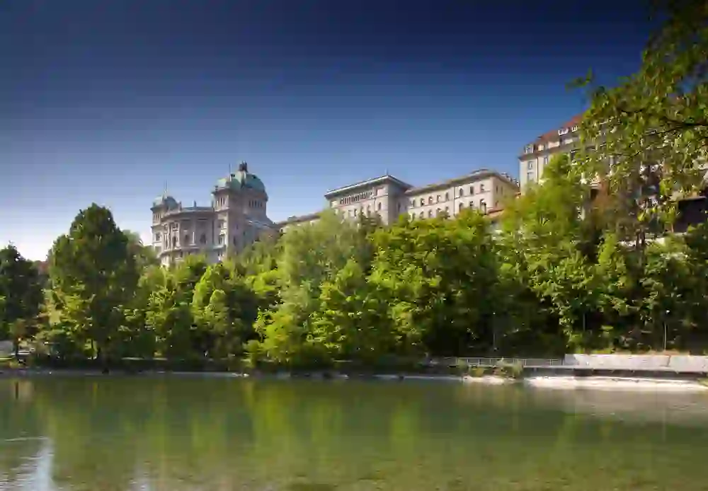 L'immagine presenta il Palazzo Federale di Berna, enfatizzato dalle acque scintillanti dell'Aar e dal verde fresco degli alberi, ed emana un'atmosfera pacifica e maestosa.