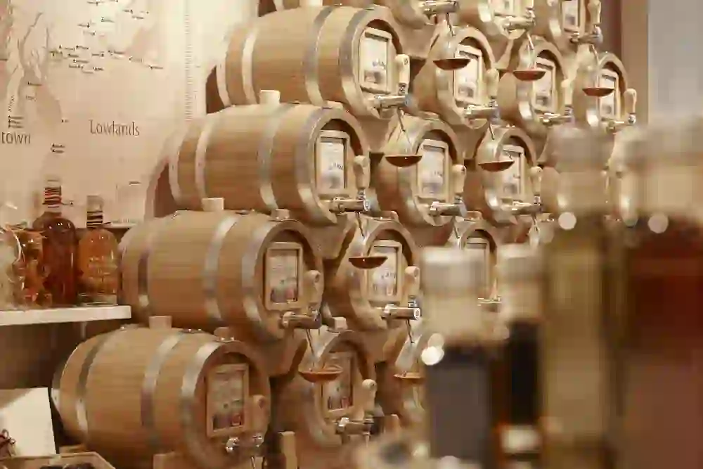Das Bild zeigt eine Auswahl an Holzfässern in einem Geschäft, vermutlich einem "Vom Fass" Laden, wo hochwertige Spirituosen, Öle, Essige und andere Flüssigkeiten zum Ausschank bereitstehen. Jedes Fass ist mit einem Hahn versehen und trägt ein Etikett, das Auskunft über den Inhalt gibt. Im Hintergrund ist eine Karte zu erkennen, die möglicherweise die Herkunftsregionen der Produkte zeigt. Das Geschäft bietet eine warme, einladige Atmosphäre für Feinschmecker.