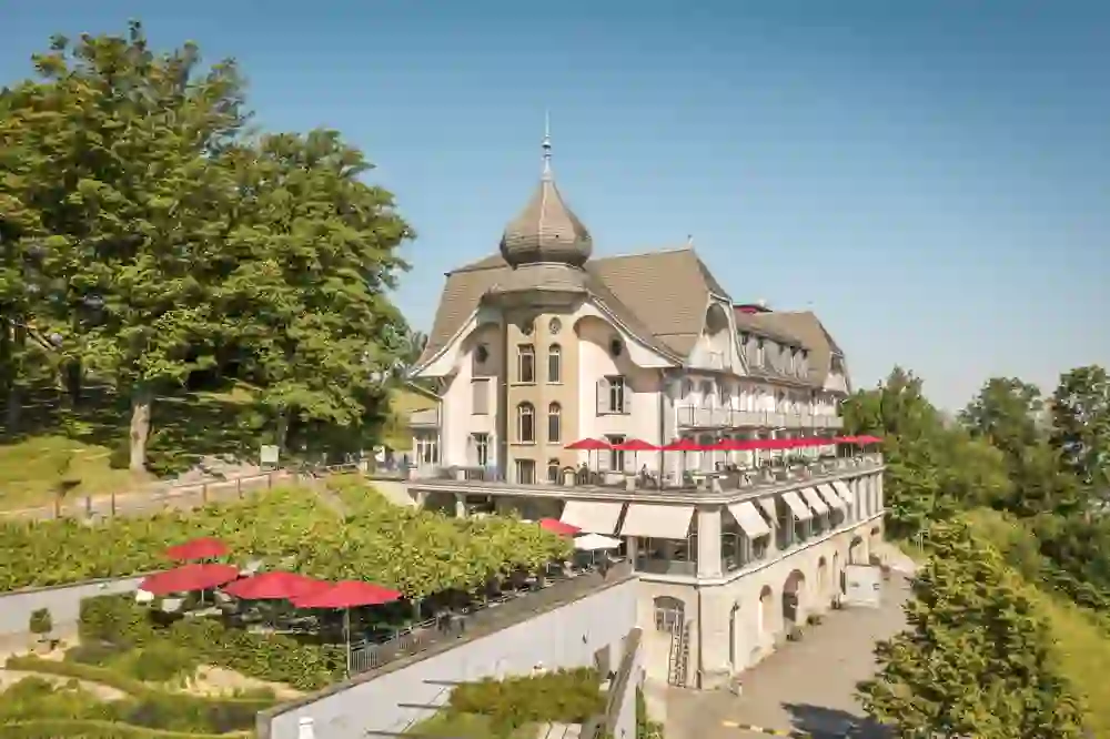 Un bâtiment majestueux avec une terrasse spacieuse, entourée d'arbres luxuriants, rayonne sous un ciel clair. Les parasols rouges ajoutent une touche colorée qui invite à se détendre dans un environnement calme et ensoleillé.