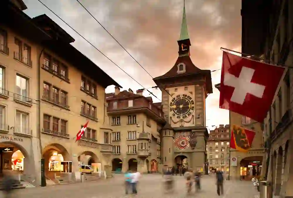 The picture shows the Zytglogge, a landmark of the city of Bern in Switzerland. It is a medieval tower with a large clock that still works. A large Swiss flag flies in front of the tower and a few people can be seen on the street.