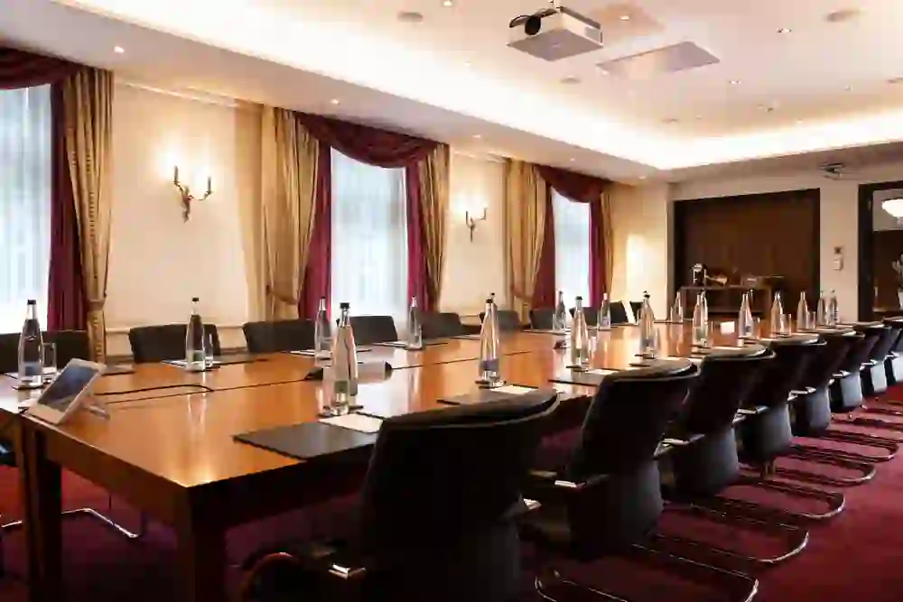 Das Bild zeigt einen stilvollen Konferenzraum mit einem großen, polierten Holztisch, umgeben von eleganten schwarzen Ledersesseln. Die Vorhänge in kräftigem Rot und Gold sowie die Wandleuchten verleihen dem Raum eine edle und professionelle Atmosphäre. Ideal für wichtige Geschäftsbesprechungen oder Präsentationen.