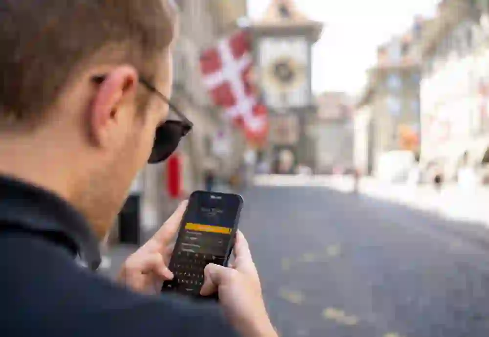 L'image montre un homme qui utilise un smartphone. En arrière-plan, on distingue de manière floue une rue historique avec des drapeaux suisses et la fameuse tour Zytglogge de Berne. L'atmosphère semble détendue et focalisée.