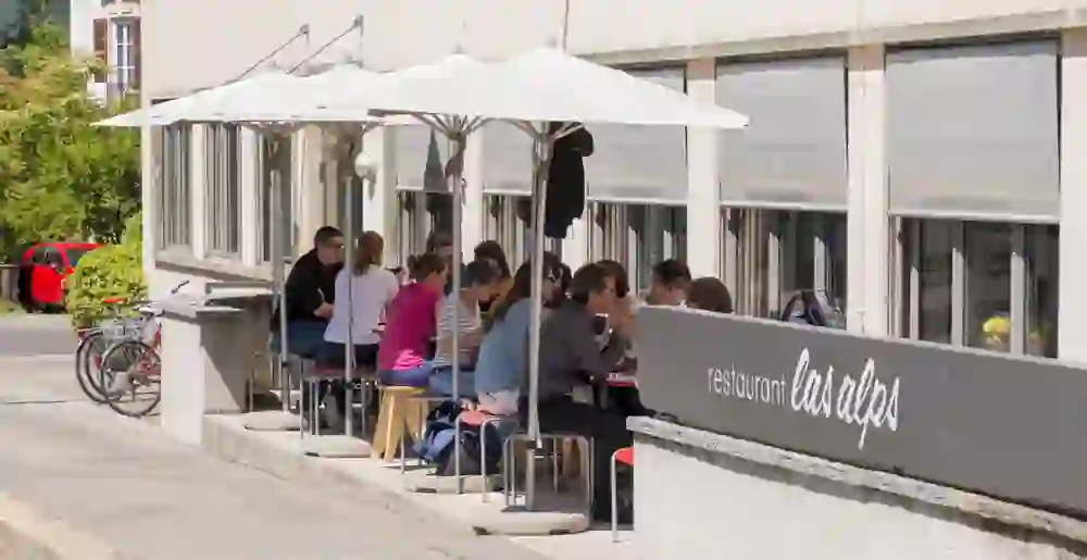 Das Bild zeigt Gäste, die in einem Außenbereich eines Restaurants namens "las alps" sitzen und essen, begleitet von sonnigem Wetter und einer entspannten, geselligen Stimmung.