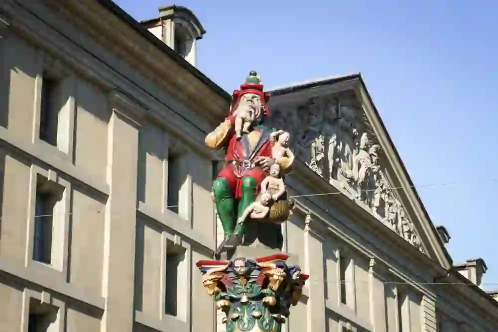 The picture shows another view of the Kindlifresserbrunnen fountain on Kornhausplatz in Bern. The fountain figure is enthroned on a column and the basin of the fountain can be seen below it. Part of the city's classical sandstone architecture is recognisable in the background.
