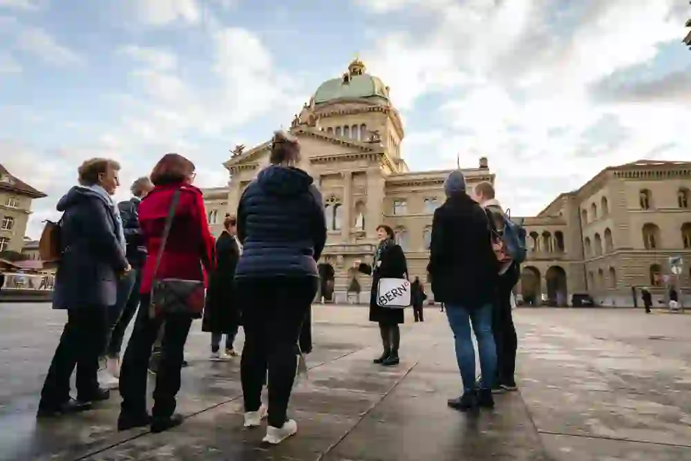 L'immagine mostra un gruppo di persone che ascoltano una guida della città sulla Bundesplatz, di fronte al Bundeshaus di Berna. Il sole proietta ombre morbide e crea un'atmosfera rilassata e attenta, mentre gli ascoltatori, avvolti in abiti caldi, appaiono interessati e curiosi. L'atmosfera è tranquilla e culturalmente arricchente.