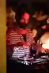 Auf dem Bild ist ein DJ zu sehen, der bei einem Event in einer Location auflegt. Er hat langes, lockiges Haar und trägt einen Kopfhörer.