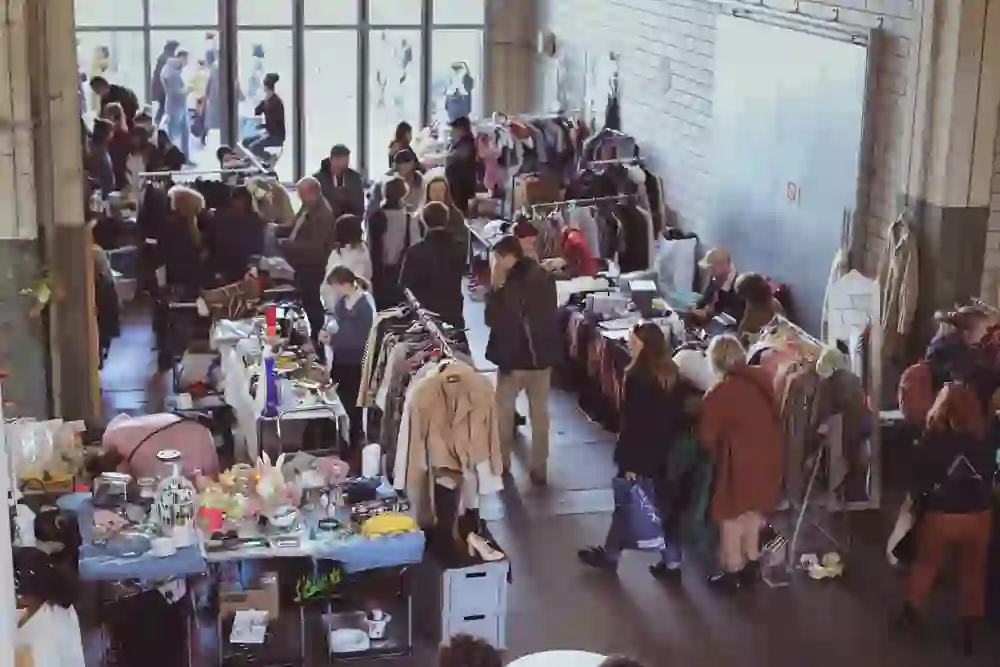 The photo shows the lively scene at the Stage flea market in Bern with people browsing between the many different stalls. The atmosphere is characterised by a mixture of curiosity and the joy of discovery, while rays of light fall through large windows and bathe the finds in warm light.
