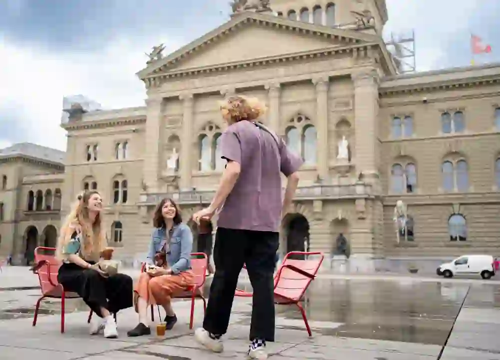 Zwei lachende Frauen geniessen in roten Stühlen eine Pause auf dem Bundesplatz in Bern, während ein Mann ihnen gegenübersteht. Die entspannte Stimmung kontrastiert mit dem ernsten Hintergrund des Bundeshauses, ein Spiel von Alltag und Geschichte.