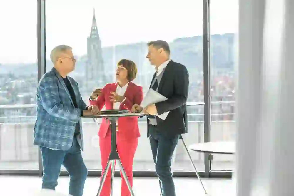 Three people have an intense conversation in a bright, modern room with a panoramic view of the city of Bern, exchanging ideas - a scene full of energy and professional dynamism.