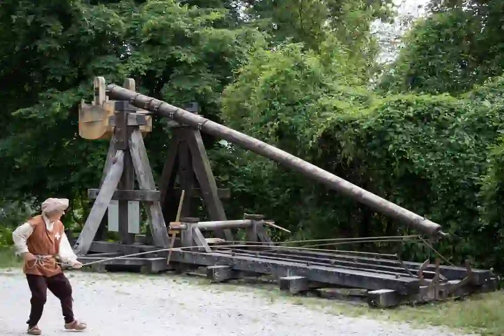 Auf dem Bild ist ein Mann in historischer Kleidung zu sehen, der neben einer grossen, alten Belagerungswaffe steht, die wie ein Katapult oder eine Kanone aussieht.