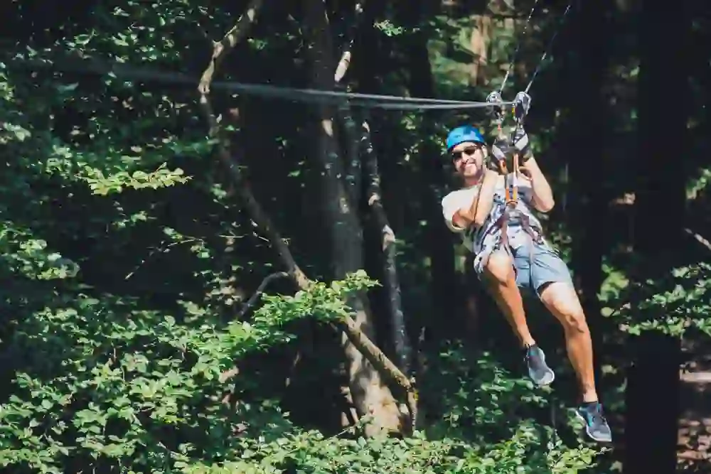 A smiling man in the rope park swings with helmet and safety equipment on a zipline through the living green of the trees, an expression of joy and freedom.