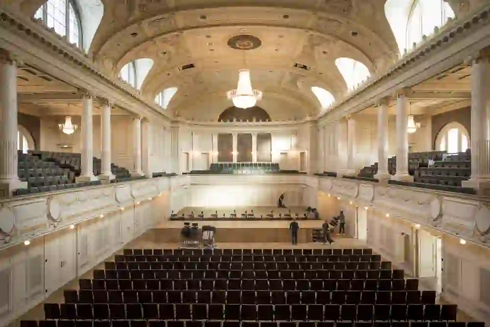 L'image montre l'intérieur élégant d'une salle de concert du Casino de Berne à l'architecture classique. Une lumière chaude éclaire les décorations artistiques et crée une atmosphère d'anticipation et de respect, tandis que des personnes se préparent sur scène pour un événement à venir.