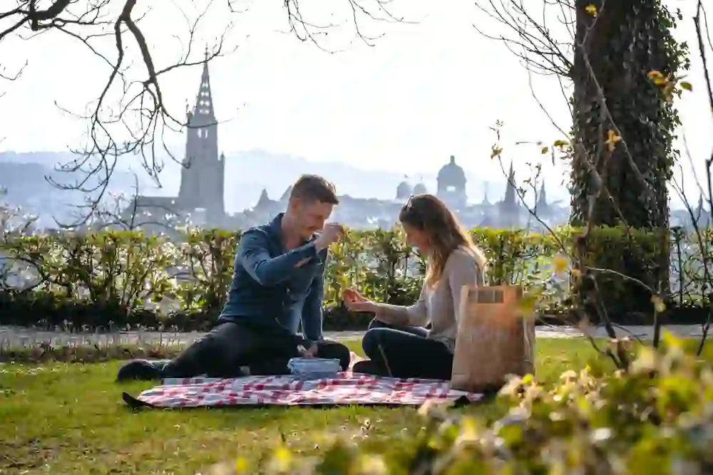 La photo montre un couple en train de pique-niquer sur une prairie avec une couverture à carreaux rouges et blancs. Ils profitent du temps ensemble et semblent échanger des œufs de Pâques ou des petits en-cas. En arrière-plan, on aperçoit une ville avec des bâtiments remarquables et des clochers d'église, ce qui pourrait faire penser à Berne, la capitale de la Suisse. Un sac en papier portant l'inscription "BERN" renforce cette impression. Le décor est empreint d'une atmosphère chaleureuse et agréable, avec des plantes en fleurs et la lumière du soleil qui illumine la scène.
