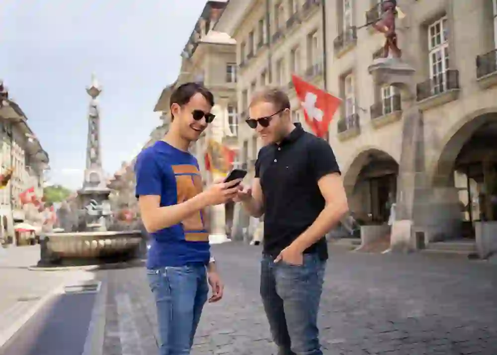 Auf dem Bild sehen wir zwei Männer in lässiger Kleidung, die gemeinsam auf ein Smartphone blicken und lächeln. Im Hintergrund erstreckt sich eine belebte Straße in Bern mit traditioneller Architektur und einer Schweizer Flagge, was eine entspannte und freundliche Atmosphäre vermittelt.