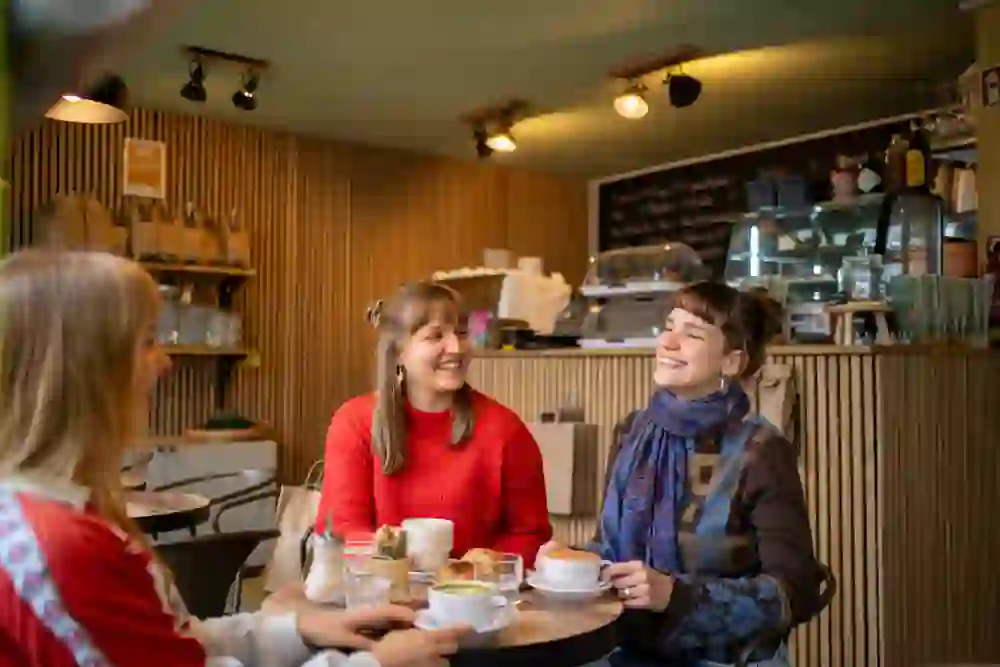 Auf dem Foto sieht man drei Personen, die in einem Café sitzen und sich unterhalten. Zwei Frauen lächeln sich an, während eine Dritte ihnen gegenüber sitzt. Auf dem Tisch stehen Getränke und Snacks, und im Hintergrund ist die Theke des Cafés mit Kaffeezubehör und weiteren Artikeln zu erkennen. Die Einrichtung ist modern und gemütlich.