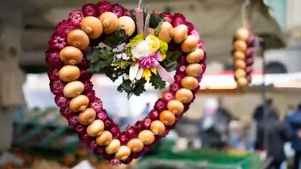 Ein bunter Zwiebelkranz schmückt das Bild, kunstvoll mit herbstlichen Blumen verziert, ein Symbol des traditionellen Zibelemärit in Bern, der lebhaften und herzlichen Atmosphäre des Marktes inmitten des städtischen Trubels.