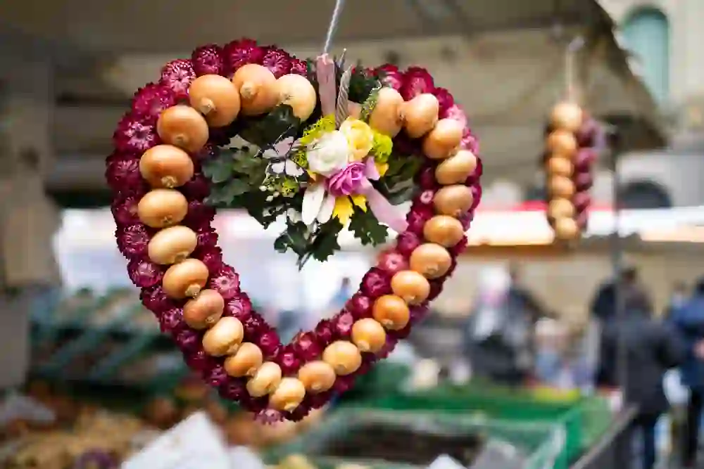 Ein bunter Zwiebelkranz schmückt das Bild, kunstvoll mit herbstlichen Blumen verziert, ein Symbol des traditionellen Zibelemärit in Bern, der lebhaften und herzlichen Atmosphäre des Marktes inmitten des städtischen Trubels.