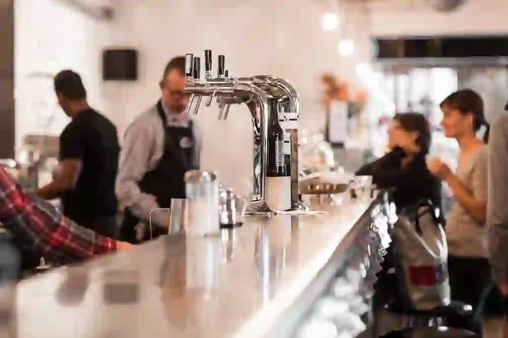 L'image capture l'atmosphère animée d'un café-bar, avec des clients en arrière-plan et un robinet brillant au centre, suggérant la convivialité.
